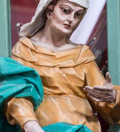 Zdjęcie nr 5: Rzeźba pełna, ustawiona na niewielkim cokole. Figura ukazana w całej postaci, zwrócona w trzech czwartych w lewo, w silnym kontrapoście, o esowato wygiętej sylwetce. Głowa delikatnie pochylona, skierowana w lewo; lewa ręka ugięta i uniesiona, prawa opuszczona z przodu, podtrzymuje płaszcz. Twarz szczupła, o wyraźnie zaznaczonej linii brody i kości policzkowych, oczy duże, płytko rzeźbione, nos niewielki. Święta jest ubrana w sięgającą ziemi,  żółtą suknię, przepasaną w talii, z wąskimi rękawami oraz dużym dekoltem z wywiniętą partią draperii przy brzegu. Tkanina przylega do ciała, jest marszczona w partii torsu i drapowana dołem w grube, pojedyncze fałdy. Turkusowy płaszcz przerzucony przez prawe przedramię opada przez plecy na lewy bok, z podtrzymywaną z przodu partią tkaniny, draperia silnie fałdowana. Na głowie długi, biały welon opadający na plecy. Polichromia naturalistyczna.
