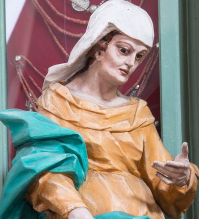 Zdjęcie nr 1: Rzeźba pełna, ustawiona na niewielkim cokole. Figura ukazana w całej postaci, zwrócona w trzech czwartych w lewo, w silnym kontrapoście, o esowato wygiętej sylwetce. Głowa delikatnie pochylona, skierowana w lewo; lewa ręka ugięta i uniesiona, prawa opuszczona z przodu, podtrzymuje płaszcz. Twarz szczupła, o wyraźnie zaznaczonej linii brody i kości policzkowych, oczy duże, płytko rzeźbione, nos niewielki. Święta jest ubrana w sięgającą ziemi,  żółtą suknię, przepasaną w talii, z wąskimi rękawami oraz dużym dekoltem z wywiniętą partią draperii przy brzegu. Tkanina przylega do ciała, jest marszczona w partii torsu i drapowana dołem w grube, pojedyncze fałdy. Turkusowy płaszcz przerzucony przez prawe przedramię opada przez plecy na lewy bok, z podtrzymywaną z przodu partią tkaniny, draperia silnie fałdowana. Na głowie długi, biały welon opadający na plecy. Polichromia naturalistyczna.
