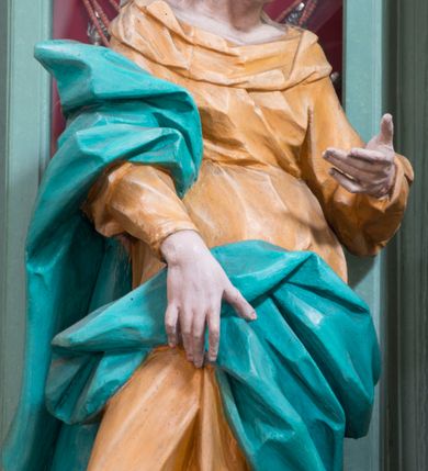 Zdjęcie nr 4: Rzeźba pełna, ustawiona na niewielkim cokole. Figura ukazana w całej postaci, zwrócona w trzech czwartych w lewo, w silnym kontrapoście, o esowato wygiętej sylwetce. Głowa delikatnie pochylona, skierowana w lewo; lewa ręka ugięta i uniesiona, prawa opuszczona z przodu, podtrzymuje płaszcz. Twarz szczupła, o wyraźnie zaznaczonej linii brody i kości policzkowych, oczy duże, płytko rzeźbione, nos niewielki. Święta jest ubrana w sięgającą ziemi,  żółtą suknię, przepasaną w talii, z wąskimi rękawami oraz dużym dekoltem z wywiniętą partią draperii przy brzegu. Tkanina przylega do ciała, jest marszczona w partii torsu i drapowana dołem w grube, pojedyncze fałdy. Turkusowy płaszcz przerzucony przez prawe przedramię opada przez plecy na lewy bok, z podtrzymywaną z przodu partią tkaniny, draperia silnie fałdowana. Na głowie długi, biały welon opadający na plecy. Polichromia naturalistyczna.
