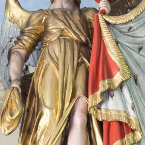 Zdjęcie nr 1: Ustawiona na niewysokim cokole pełnoplastyczna figura anioła przytrzymującego baldachim w zwieńczeniu ołtarza głównego. Anioł spogląda w dół, stoi w lekkim kontrapoście z lewą nogą wysuniętą do przodu; lewą ręką przytrzymuje materiał baldachimu, prawą fragment swojego płaszcza. Ma owalną, pełną twarz, małe oczy, zadarty nos oraz niewielkie usta. Długie, jasnobrązowe, kręcone włosy spływają mu na plecy. Ubrany jest w długą, złotą suknię przewiązaną w pasie i spływającą w dół wąskimi fałdami; rękawy podciągnięte; na nogach sandały sięgające do połowy łydki. Skrzydła rozłożone, w górnych partiach złocone. 