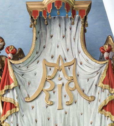 Zdjęcie nr 3: Ustawiona na niewysokim cokole pełnoplastyczna figura anioła przytrzymującego baldachim w zwieńczeniu ołtarza głównego. Anioł spogląda w dół, stoi w lekkim kontrapoście z lewą nogą wysuniętą do przodu; lewą ręką przytrzymuje materiał baldachimu, prawą fragment swojego płaszcza. Ma owalną, pełną twarz, małe oczy, zadarty nos oraz niewielkie usta. Długie, jasnobrązowe, kręcone włosy spływają mu na plecy. Ubrany jest w długą, złotą suknię przewiązaną w pasie i spływającą w dół wąskimi fałdami; rękawy podciągnięte; na nogach sandały sięgające do połowy łydki. Skrzydła rozłożone, w górnych partiach złocone. 