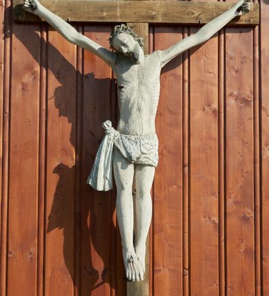 Zdjęcie nr 1: Figura Chrystusa na prostym krzyżu umieszczona na zewnątrz kościoła. Rzeźba Chrystusa przybita do krzyża trzema gwoździami, ukazana w typie Cristo morto. Ciało w głębokim zwisie, sylwetka wydłużona o delikatnie podkreślonej anatomii, nogi lekko ugięte w kolanach, stopy założone prawa na lewą. Chrystus ma głowę przechyloną na prawe ramię. Twarz szczupła z wąskim i długim nosem, zamkniętymi oczami, rozchylonymi ustami, okolona krótką brodą. Włosy długie, spływające na prawe ramię i plecy skręconymi lokami, na głowie założona korona cierniowa. Perizonium zawiązane na węzeł na prawym boku, z jednym końcem opadającym wzdłuż prawej nogi. Z boku, dłoni, stóp i spod korony cierniowej spływają strużki krwi. Rzeźba prawie w całości polichromowana na biało, włosy i broda na brązowo. Krzyż i titulus nowe, proste, titulus z napisem „INRI.”. 
