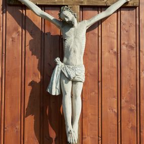 Zdjęcie nr 1: Figura Chrystusa na prostym krzyżu umieszczona na zewnątrz kościoła. Rzeźba Chrystusa przybita do krzyża trzema gwoździami, ukazana w typie Cristo morto. Ciało w głębokim zwisie, sylwetka wydłużona o delikatnie podkreślonej anatomii, nogi lekko ugięte w kolanach, stopy założone prawa na lewą. Chrystus ma głowę przechyloną na prawe ramię. Twarz szczupła z wąskim i długim nosem, zamkniętymi oczami, rozchylonymi ustami, okolona krótką brodą. Włosy długie, spływające na prawe ramię i plecy skręconymi lokami, na głowie założona korona cierniowa. Perizonium zawiązane na węzeł na prawym boku, z jednym końcem opadającym wzdłuż prawej nogi. Z boku, dłoni, stóp i spod korony cierniowej spływają strużki krwi. Rzeźba prawie w całości polichromowana na biało, włosy i broda na brązowo. Krzyż i titulus nowe, proste, titulus z napisem „INRI.”. 
