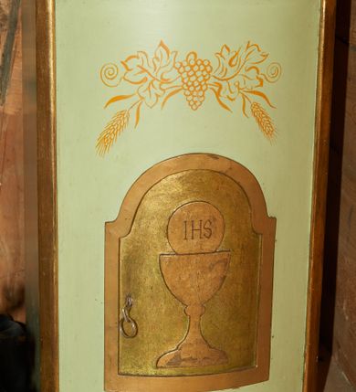 Zdjęcie nr 1: Tabernakulum w formie prostopadłościennej szafki z lekko wypukłą przednią ścianką, ustawione na niskim cokoliku i zamknięte prostym gzymsem. Drzwiczki prostokątne zamknięte łukiem nadwieszanym z płaskorzeźbionym przedstawieniem kielicha z hostią w polu. Boki i drzwiczki tabernakulum złocone, front w kolorze jasnoszarym, nad drzwiczkami malowane w kolorze złotym winne grona i kłosy. 