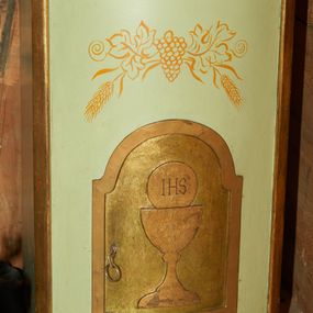 Zdjęcie nr 1: Tabernakulum w formie prostopadłościennej szafki z lekko wypukłą przednią ścianką, ustawione na niskim cokoliku i zamknięte prostym gzymsem. Drzwiczki prostokątne zamknięte łukiem nadwieszanym z płaskorzeźbionym przedstawieniem kielicha z hostią w polu. Boki i drzwiczki tabernakulum złocone, front w kolorze jasnoszarym, nad drzwiczkami malowane w kolorze złotym winne grona i kłosy. 