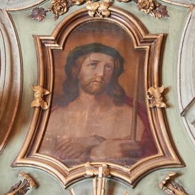 Zdjęcie nr 1: Obraz w kształcie stojącego prostokąta umieszczony w ramie o wklęsłych bokach, potrójnie wklęsłej podstawie, zwieńczonej łukiem nadwieszonym. Chrystus w typie Ecce Homo, ukazany w popiersiu, frontalnie z głową przechyloną na lewą stronę i delikatnie uniesioną do góry. Zbawiciel ma ręce skrzyżowane na piersi i związane sznurem, w prawej dłoni trzyma trzcinę. Twarz o idealizowanych rysach z długim nosem, dużymi oczami ze wzrokiem skierowanym ku górze, okolona krótką brodą oraz długimi, bujnymi i brązowymi włosami. Chrystus ubrany jest w czerwony płaszcz, założony na ramiona i odsłaniający nagi tors; na głowie ma założoną obszerną, zieloną koronę cierniową. Tło jednolite, ugrowe.