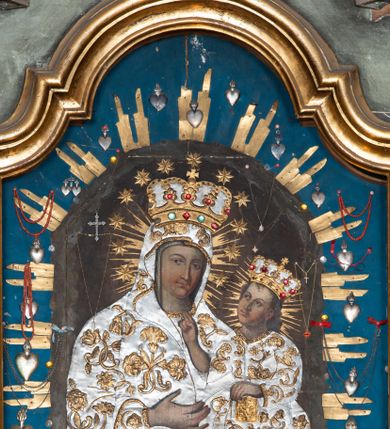 Zdjęcie nr 1: Obraz w kształcie stojącego prostokąta zamknięty łukiem wklęsło-wypukłym z uskokami, przedstawiający Matkę Boską z Dzieciątkiem. Maria została ukazana w półpostaci z Dzieciątkiem Jezus na lewym ręku, z jabłkiem w prawej dłoni (przysłoniętym sukienką), z głową delikatnie przechyloną na lewe ramię i wzrokiem skierowanym na widza. Twarz Marii o delikatnych rysach z drobnymi ustami, długim nosem i zarumienionymi policzkami. Dzieciątko ukazane jest w pozycji siedzącej, zwrócone trzy czwarte w prawo, prawą rączką czyni gest błogosławieństwa, lewą opiera na księdze. Twarz Dzieciątka owalna, pełna z długim nosem i półkolistymi łukami brwiowymi, policzki silnie zarumienione. Na postacie Marii i Dzieciątka nałożone są metalowe, złocone i srebrzone sukienki, dekorowane w motywy floralne, a także korony w typie zamkniętym. Karnacje blade. Całość ukazana jest na ciemnym tle, w którym wyróżniają się promieniste, złocone nimby wokół głów Marii i Dzieciątka. Obraz umieszczony na niebieskim tle, obwiedzionym wiązkami metalowych, złoconych promieni oraz zawieszonych wotów. Rama drewniana, profilowana złocona.