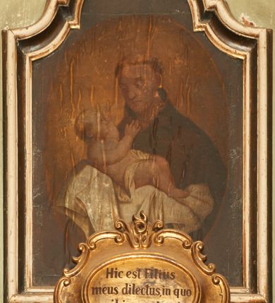 Zdjęcie nr 1: Obraz w kształcie stojącego prostokąta, umieszczony w drewnianej ramie zamkniętej łukiem wklęsło-wypukłym z uskokami. Kompozycja ujęta w owalu, na ugrowym tle. W centrum ukazany jest św. Antoni Padewski w półpostaci, trzy czwarte zwrócony w prawo z Dzieciątkiem Jezus na rękach. Twarz okrągła z drobnymi ustami, dużym nosem i przymkniętymi oczami. Święty ubrany jest w habit franciszkański, na głowie ma tonsurę. Dzieciątko ukazane jest w pozycji półleżącej z rączkami wyciągniętymi w stronę twarzy świętego, ułożone na białej tkaninie i przewiązane w biodrach pieluszką. Tkanina obwiedziona jest na brzegach delikatną, fantazyjną bordiurą. Twarz o dziecięcych rysach, włosy jasne i krótkie, wokół głowy promienisty nimb. 