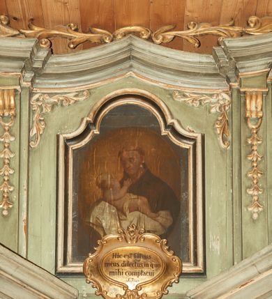 Zdjęcie nr 2: Obraz w kształcie stojącego prostokąta, umieszczony w drewnianej ramie zamkniętej łukiem wklęsło-wypukłym z uskokami. Kompozycja ujęta w owalu, na ugrowym tle. W centrum ukazany jest św. Antoni Padewski w półpostaci, trzy czwarte zwrócony w prawo z Dzieciątkiem Jezus na rękach. Twarz okrągła z drobnymi ustami, dużym nosem i przymkniętymi oczami. Święty ubrany jest w habit franciszkański, na głowie ma tonsurę. Dzieciątko ukazane jest w pozycji półleżącej z rączkami wyciągniętymi w stronę twarzy świętego, ułożone na białej tkaninie i przewiązane w biodrach pieluszką. Tkanina obwiedziona jest na brzegach delikatną, fantazyjną bordiurą. Twarz o dziecięcych rysach, włosy jasne i krótkie, wokół głowy promienisty nimb. 