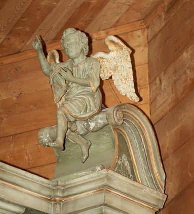 Zdjęcie nr 1: Dwie pełnoplastyczne figury siedzących na odcinkach przyczółka aniołów. Aniołek po prawej stronie wznosi prawą rękę ku górze, a lewą trzyma na wysokości piersi, nogi ukazane w ruchu, prawa noga wyciągniętą do przodu. Aniołek po lewej stronie ołtarza obie ręce wyciąga w swoją lewą stronę, a stopy ma skrzyżowane, kolana rozchylone na boki. Twarze pełne z dużymi ustami i krótkimi nosami, delikatnie zaznaczonymi oczami; włosy krótkie, ułożone w grube pukle. Postacie ubrane są w krótkie tuniki oraz zarzucone  na plecy płaszcze, szaty obwiedzione są złotą lamówką. Na plecach anioły mają duże, rozłożone skrzydła. Rzeźby w całości polichromowane są na kolor biały, skrzydła złocone. 