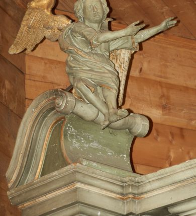 Zdjęcie nr 2: Dwie pełnoplastyczne figury siedzących na odcinkach przyczółka aniołów. Aniołek po prawej stronie wznosi prawą rękę ku górze, a lewą trzyma na wysokości piersi, nogi ukazane w ruchu, prawa noga wyciągniętą do przodu. Aniołek po lewej stronie ołtarza obie ręce wyciąga w swoją lewą stronę, a stopy ma skrzyżowane, kolana rozchylone na boki. Twarze pełne z dużymi ustami i krótkimi nosami, delikatnie zaznaczonymi oczami; włosy krótkie, ułożone w grube pukle. Postacie ubrane są w krótkie tuniki oraz zarzucone  na plecy płaszcze, szaty obwiedzione są złotą lamówką. Na plecach anioły mają duże, rozłożone skrzydła. Rzeźby w całości polichromowane są na kolor biały, skrzydła złocone. 