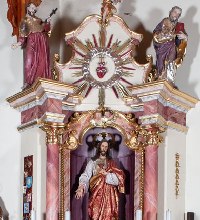Zdjęcie nr 1: Ołtarz przyścienny, architektoniczny, jednoosiowy, jednokondygnacyjny. Mensa drewniana o formach wklęsło-wypukłych; na licu płycina w formie leżącego prostokąta o ściętych narożach wypełniona marmoryzacją. Tabernakulum dwudrzwiczkowe, zamknięte łukiem pełnym, ujęte dwiema  kolumienkami o kompozytowych kapitelach. W zwieńczeniu ażurowa, złocona dekoracja w formie przeciwstawnie ustawionego akantu z palmetą na osi. Na drzwiczkach płaskorzeźbione kłosy oraz winne grona. Retabulum na wysokim cokole ujęte ustawionymi ukośnie kolumnami o kompozytowych kapitelach i marmoryzowanych trzonach umiejscowionymi na wysokich postumentach (lica zdobione marmoryzowanymi płycinami  wypełnioymi rocaille&#039;m). Podpory dźwigają odcinki pełnego, wyłamanego belkowania zwieńczonego wydatnym, silnie profilowanym gzymsem. Na belkowaniu, na osiach kolumn pełnoplastyczne figury niezidentyfikowanych świętych z krzyżem w ręku (z lewej, św. Jan Nepomucen?) oraz z księgą (z prawej). W polu centralnym nisza zamknięta u góry łukiem o falistym zarysie utworzonym przez dwie przeciwstawnie ustawione woluty. Nisza mieści pełnoplastyczną figurę Chrystusa z gorejącym sercem. Ponad nią, a osi dwie płaskorzeźbione uskrzydlone główki anielskie. Zwieńczenie o wolutowo wyciętych bokach zamknięte łukiem wklęsło-wypukłym. W polu płaskorzeźbione przedstawienie gorejącego serca w srebrzonym medalionie z obłoków oraz glorii promienistej o alternowanych promieniach. Powyżej na osi, pełnoplastyczna figura uskrzydlonego anioła. Po bokach wota i kampanule. Struktura malowana na kolor jasnobeżowy. Marmoryzacja w odcieniach różu. Ornamentyka oraz załamania profili architektonicznych złocone.
