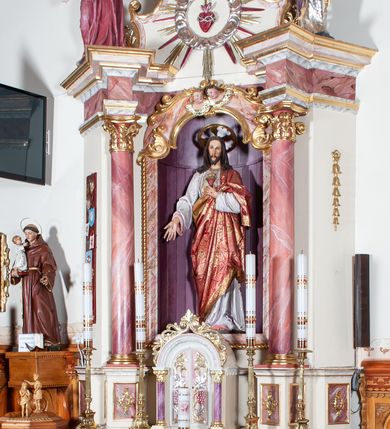 Zdjęcie nr 2: Ołtarz przyścienny, architektoniczny, jednoosiowy, jednokondygnacyjny. Mensa drewniana o formach wklęsło-wypukłych; na licu płycina w formie leżącego prostokąta o ściętych narożach wypełniona marmoryzacją. Tabernakulum dwudrzwiczkowe, zamknięte łukiem pełnym, ujęte dwiema  kolumienkami o kompozytowych kapitelach. W zwieńczeniu ażurowa, złocona dekoracja w formie przeciwstawnie ustawionego akantu z palmetą na osi. Na drzwiczkach płaskorzeźbione kłosy oraz winne grona. Retabulum na wysokim cokole ujęte ustawionymi ukośnie kolumnami o kompozytowych kapitelach i marmoryzowanych trzonach umiejscowionymi na wysokich postumentach (lica zdobione marmoryzowanymi płycinami  wypełnioymi rocaille'm). Podpory dźwigają odcinki pełnego, wyłamanego belkowania zwieńczonego wydatnym, silnie profilowanym gzymsem. Na belkowaniu, na osiach kolumn pełnoplastyczne figury niezidentyfikowanych świętych z krzyżem w ręku (z lewej, św. Jan Nepomucen?) oraz z księgą (z prawej). W polu centralnym nisza zamknięta u góry łukiem o falistym zarysie utworzonym przez dwie przeciwstawnie ustawione woluty. Nisza mieści pełnoplastyczną figurę Chrystusa z gorejącym sercem. Ponad nią, a osi dwie płaskorzeźbione uskrzydlone główki anielskie. Zwieńczenie o wolutowo wyciętych bokach zamknięte łukiem wklęsło-wypukłym. W polu płaskorzeźbione przedstawienie gorejącego serca w srebrzonym medalionie z obłoków oraz glorii promienistej o alternowanych promieniach. Powyżej na osi, pełnoplastyczna figura uskrzydlonego anioła. Po bokach wota i kampanule. Struktura malowana na kolor jasnobeżowy. Marmoryzacja w odcieniach różu. Ornamentyka oraz załamania profili architektonicznych złocone.