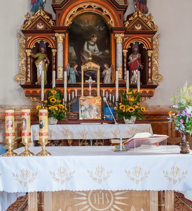 Zdjęcie nr 2: Ołtarz przyścienny, architektoniczny, trójosiowy. Mensa ołtarzowa skrzyniowa, prostopadłościenna z kamiennym portatylem, zamknięta profilowanym gzymsem podtrzymywanym przez cztery marmoryzowane kolumny. Antepedium dekorowane trzema płycinami: środkowa kwadratowa z krzyżem trójlistnym w czworoliściu, boczne  z aplikowanym motywem winorośli i kłosów zbóż. Retabulum ustawione na predelli, trójosiowe o półkoliście zamkniętych wnękach zakończonych trójkątnymi szczytami, w polu których uskrzydlone główki anielskie w medalionach. Wnęka środkowa wyższa i szersza zamknięta dwuspadowym daszkiem, ujęta parą kolumn kompozytowych wspierających odcinki gzymsu. Wewnątrz obraz z przedstawieniem  św. Rozalii. We wnękach bocznych pełnoplastyczne figury św. Piotra po lewej i św. Pawła po prawej stronie.  Nad wnęką środkową baldachim z figurą Chrystusa Zmartwychwstałego, po bokach pełnoplastyczne figury świętych. Predella dekorowana fryzem arkadkowym oraz płycinami z maswerkowaniem i rozetami, boki retabulum ujęte ażurowymi uszakami formowanymi z wici roślinnej. Półkoliste łuki wnęk zdobione arkadkami. Całość polichromowana na odcień ciemnobrązowy, detal architektoniczny i profilowania pomarańczowe oraz złocone. 