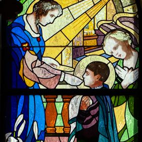 Zdjęcie nr 1: Witraż w kształcie stojącego prostokąta zamkniętego półkoliście z przedstawieniem św. Stanisława Kostki. Święty ukazany jako młody chłopiec, z lewego profilu, w pozycji klęczącej, w momencie przyjmowania Komunii Świętej z rąk stojącego naprzeciw niego Anioła. Ubrany jest w strój jezuity i niebieski płaszcz spływający na ziemię. Anioł udzielający Komunii ubrany jest w spodnią, bladoróżową szatę z długimi rękawami oraz wierzchnią ciemnoniebieską, z szerokimi, rozciętymi rękawami. Za postacią świętego widoczny drugi anioł ze złożonymi na piersiach rękami, ubrany w szatę w odcieniach zieleni. Scena ukazana na tle sumarycznie oddanej przestrzeni architektonicznej. U dołu, po lewej stronie gałązki białych lilii. W górnej partii witrażu łuk z malowaną dekoracją ornamentalną i snop światła padający na anioła. W dolnej części napisy, od lewej: „FUNDOWALI: / IGNACY I / JÓZEF / GAJNIAK”, pośrodku „św(ięty) STANISŁAWIE KOSTKO / MÓDL SIĘ ZA NAMI”, po prawej „JABŁONKA / ORAWSKA / 1938”.
