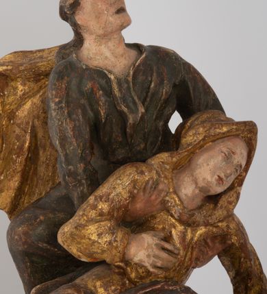 Zdjęcie nr 6: Rzeźbiarska grupa Ukrzyżowania z Matką Boską, św. Janem Ewangelistą i św. Marią Magdaleną ustawiona na cokole, wspartym na dwóch wolutowych nóżkach i trzeciej w formie liścia. Frontalna ścianka cokołu flankowana pilastrami i zamknięta gzymsem utworzonym z dwóch wolut, spiętych pośrodku kulą, dekorowanym perełkowaniem. Jego pole zdobione jest esownicami, palmami oraz niewielką rzeźbą węża gryzącego jabłko po lewej i czaszką po prawej stronie cokołu. Na cokole ustawiona grupa rzeźbiarska: nieco z prawej strony krucyfiks i św. Maria Magdalena, po lewej św. Jan Ewangelista i Matka Boska. Chrystus przybity do krzyża trzema gwoździami. Figura w dużym zwisie o układzie rąk bliskim literze „V”, sylwetka esowato wygięta, stopy ułożone w układzie prawa na lewą, głowa opuszczona na prawe ramię. Twarz okolona zarostem, włosy długie, spływające na prawe ramię, na głowie korona cierniowa oraz krzyżowy nimb promienisty, zachowany fragmentarycznie. Ciało wychudzone o plastycznie kształtowanej anatomii z podkreśloną muskulaturą i zaznaczonymi śladami męki. Perizonium w formie tkaniny przewiązanej sznurem, z odsłoniętym prawym biodrem oraz zwisem z tyłu. Krzyż gładki, pomalowany na czarno. Poniżej figura św. Marii Magdaleny ukazana w pozycji klęczącej, rękami obejmująca krzyż, ustawiona na niskim cokole. Święta widoczna z lewego profilu. Twarz szeroka, usta małe, oczy zamknięte, czoło wysokie, długie włosy opadają na plecy. Święta ubrana w długą suknię z krótkimi rękawami oraz obszerny płaszcz okrywający ją z prawego boku, którego jedną połę trzyma w dłoniach. Po lewej stronie kompozycji św. Jan Ewangelista podtrzymujący rękami mdlejącą Matkę Boską. Święty zwrócony delikatnie w lewo, z głową skierowaną w stronę krzyża. Twarz szeroka, nos długi i szeroki, oczy skierowane ku górze, usta wąskie, długie włosy opadają na plecy. Święty ubrany w długą suknię z długimi rękawami oraz płaszcz rozwiany za jego plecami. Poniżej Matka Boska, która opada bezwładnie, prawą rękę trzyma na piersi, lewą opuszcza w dół. Twarz szeroka z zamkniętymi oczami i małymi ustami. Ubrana w długą suknię z długimi rękawami, welon na głowie oraz płaszcz otulający nogi. Polichromia naturalistyczna w odsłoniętych partiach ciała, szaty i ornamenty na cokole złocone.
