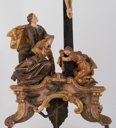 Zdjęcie nr 1: Rzeźbiarska grupa Ukrzyżowania z Matką Boską, św. Janem Ewangelistą i św. Marią Magdaleną ustawiona na cokole, wspartym na dwóch wolutowych nóżkach i trzeciej w formie liścia. Frontalna ścianka cokołu flankowana pilastrami i zamknięta gzymsem utworzonym z dwóch wolut, spiętych pośrodku kulą, dekorowanym perełkowaniem. Jego pole zdobione jest esownicami, palmami oraz niewielką rzeźbą węża gryzącego jabłko po lewej i czaszką po prawej stronie cokołu. Na cokole ustawiona grupa rzeźbiarska: nieco z prawej strony krucyfiks i św. Maria Magdalena, po lewej św. Jan Ewangelista i Matka Boska. Chrystus przybity do krzyża trzema gwoździami. Figura w dużym zwisie o układzie rąk bliskim literze „V”, sylwetka esowato wygięta, stopy ułożone w układzie prawa na lewą, głowa opuszczona na prawe ramię. Twarz okolona zarostem, włosy długie, spływające na prawe ramię, na głowie korona cierniowa oraz krzyżowy nimb promienisty, zachowany fragmentarycznie. Ciało wychudzone o plastycznie kształtowanej anatomii z podkreśloną muskulaturą i zaznaczonymi śladami męki. Perizonium w formie tkaniny przewiązanej sznurem, z odsłoniętym prawym biodrem oraz zwisem z tyłu. Krzyż gładki, pomalowany na czarno. Poniżej figura św. Marii Magdaleny ukazana w pozycji klęczącej, rękami obejmująca krzyż, ustawiona na niskim cokole. Święta widoczna z lewego profilu. Twarz szeroka, usta małe, oczy zamknięte, czoło wysokie, długie włosy opadają na plecy. Święta ubrana w długą suknię z krótkimi rękawami oraz obszerny płaszcz okrywający ją z prawego boku, którego jedną połę trzyma w dłoniach. Po lewej stronie kompozycji św. Jan Ewangelista podtrzymujący rękami mdlejącą Matkę Boską. Święty zwrócony delikatnie w lewo, z głową skierowaną w stronę krzyża. Twarz szeroka, nos długi i szeroki, oczy skierowane ku górze, usta wąskie, długie włosy opadają na plecy. Święty ubrany w długą suknię z długimi rękawami oraz płaszcz rozwiany za jego plecami. Poniżej Matka Boska, która opada bezwładnie, prawą rękę trzyma na piersi, lewą opuszcza w dół. Twarz szeroka z zamkniętymi oczami i małymi ustami. Ubrana w długą suknię z długimi rękawami, welon na głowie oraz płaszcz otulający nogi. Polichromia naturalistyczna w odsłoniętych partiach ciała, szaty i ornamenty na cokole złocone.
