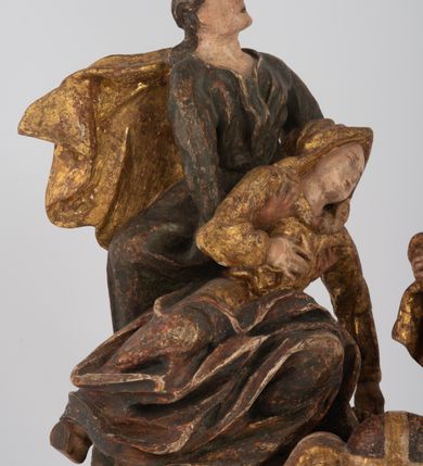 Zdjęcie nr 5: Rzeźbiarska grupa Ukrzyżowania z Matką Boską, św. Janem Ewangelistą i św. Marią Magdaleną ustawiona na cokole, wspartym na dwóch wolutowych nóżkach i trzeciej w formie liścia. Frontalna ścianka cokołu flankowana pilastrami i zamknięta gzymsem utworzonym z dwóch wolut, spiętych pośrodku kulą, dekorowanym perełkowaniem. Jego pole zdobione jest esownicami, palmami oraz niewielką rzeźbą węża gryzącego jabłko po lewej i czaszką po prawej stronie cokołu. Na cokole ustawiona grupa rzeźbiarska: nieco z prawej strony krucyfiks i św. Maria Magdalena, po lewej św. Jan Ewangelista i Matka Boska. Chrystus przybity do krzyża trzema gwoździami. Figura w dużym zwisie o układzie rąk bliskim literze „V”, sylwetka esowato wygięta, stopy ułożone w układzie prawa na lewą, głowa opuszczona na prawe ramię. Twarz okolona zarostem, włosy długie, spływające na prawe ramię, na głowie korona cierniowa oraz krzyżowy nimb promienisty, zachowany fragmentarycznie. Ciało wychudzone o plastycznie kształtowanej anatomii z podkreśloną muskulaturą i zaznaczonymi śladami męki. Perizonium w formie tkaniny przewiązanej sznurem, z odsłoniętym prawym biodrem oraz zwisem z tyłu. Krzyż gładki, pomalowany na czarno. Poniżej figura św. Marii Magdaleny ukazana w pozycji klęczącej, rękami obejmująca krzyż, ustawiona na niskim cokole. Święta widoczna z lewego profilu. Twarz szeroka, usta małe, oczy zamknięte, czoło wysokie, długie włosy opadają na plecy. Święta ubrana w długą suknię z krótkimi rękawami oraz obszerny płaszcz okrywający ją z prawego boku, którego jedną połę trzyma w dłoniach. Po lewej stronie kompozycji św. Jan Ewangelista podtrzymujący rękami mdlejącą Matkę Boską. Święty zwrócony delikatnie w lewo, z głową skierowaną w stronę krzyża. Twarz szeroka, nos długi i szeroki, oczy skierowane ku górze, usta wąskie, długie włosy opadają na plecy. Święty ubrany w długą suknię z długimi rękawami oraz płaszcz rozwiany za jego plecami. Poniżej Matka Boska, która opada bezwładnie, prawą rękę trzyma na piersi, lewą opuszcza w dół. Twarz szeroka z zamkniętymi oczami i małymi ustami. Ubrana w długą suknię z długimi rękawami, welon na głowie oraz płaszcz otulający nogi. Polichromia naturalistyczna w odsłoniętych partiach ciała, szaty i ornamenty na cokole złocone.
