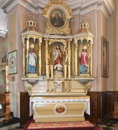 Zdjęcie nr 3: Ołtarz przyścienny, architektoniczny, trójosiowy, jednokondygnacyjny ze zwieńczeniem. Ołtarz skrzyniowy, sarkofagowy, z kamiennym portatylem. Mensa wklęsło-wypukła, z wydzielonym polem, pośrodku którego, ujęte okrągłą ramą płaskorzeźbione, polichromowane gorejące serce. Tabernakulum w formie prostopadłościennej skrzynki, ustawione na niskim cokole, ujęte parą lizen zdobionych cekinami, zamknięte profilowanych gzymsem. Drzwiczki dekorowane płyciną w kształcie stojącego prostokąta, zamkniętego łukiem półkolistym, w polu z płaskorzeźbionym barankiem siedzącym na księdze na zielonym tle, w dolnej części kompozycji srebrne obłoki, w górnej złote promienie. Retabulum ustawione na wysokim cokole, kształtowanym schodkowo, po bokach ujętym małymi spływami wolutowymi. Osie boczne retabulum w formie ażurowych, architektonicznych tempiett, o profilowanych wielobocznych podstawach z pięciobocznym podwieszeniem. Baldachimy, wsparte na parze kolumn, kształtowane są na wzór architektonicznych dachów, z wycięciem arkadkowym u dołu oraz ze zwieńczeniem w formie wielobocznej latarni zakończonej kwiatonem. W niszy po lewej figura św. Wojciecha, a po prawej św. Stanisława. Część środkowa retabulum flankowana parą kolumn wspierających wyłamane pośrodku niepełne belkowanie. W polu obraz rzeźba Najświętsze Serce Jezusa, dookoła dekorowana bujnym ornamentem akantowym. Zwieńczenie w formie owalu ujętego dekoracyjną ramą z obrazem św. Andrzeja Boboli. U szczytu rzeźbiona, uskrzydlona główka aniołka i łaciński krzyż. Polichromia w kolorze jasnobeżowym, detale zielone, profilowania i ornamentyka złocone.