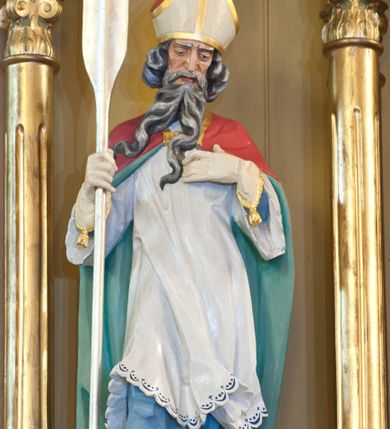 Zdjęcie nr 1: Rzeźba św. Wojciecha wydrążona z tyłu, ustawiona na niskim cokole. Święty ukazany w pozycji stojącej, frontalnie, w lekkim kontrapoście, w prawej dłoni trzyma wiosło, a lewą ma złożoną na piersi. Twarz szeroka o ostrych rysach, dużych i głęboko osadzonych oczach, długim i szerokim nosie, okolona długą, siwą i bujną brodą rozdzieloną na dwa długie pukle. Włosy średniej długości, siwe, bujne, zasłaniające uszy. Święty jest ubrany w niebieską albę, białą komżę i czerwoną kapę z jasnozieloną podszewką spiętą na piersi złotą broszą. Na dłoniach ma białe rękawiczki, na głowie biało-złotą infułę, a na stopach brązowe buty. Polichromia w odsłoniętych partiach ciała naturalistyczna, detale złocone i srebrzone.