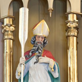 Zdjęcie nr 1: Rzeźba św. Wojciecha wydrążona z tyłu, ustawiona na niskim cokole. Święty ukazany w pozycji stojącej, frontalnie, w lekkim kontrapoście, w prawej dłoni trzyma wiosło, a lewą ma złożoną na piersi. Twarz szeroka o ostrych rysach, dużych i głęboko osadzonych oczach, długim i szerokim nosie, okolona długą, siwą i bujną brodą rozdzieloną na dwa długie pukle. Włosy średniej długości, siwe, bujne, zasłaniające uszy. Święty jest ubrany w niebieską albę, białą komżę i czerwoną kapę z jasnozieloną podszewką spiętą na piersi złotą broszą. Na dłoniach ma białe rękawiczki, na głowie biało-złotą infułę, a na stopach brązowe buty. Polichromia w odsłoniętych partiach ciała naturalistyczna, detale złocone i srebrzone.