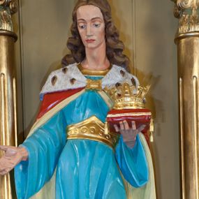 Zdjęcie nr 1: Figura św. Kazimierza Jagiellończyka ustawiona na niskim cokole. Święty ukazany w pozycji stojącej, frontalnie, w lekkim kontrapoście, w lewej dłoni trzyma koronę otwartą ułożoną na czerwonej poduszce, prawą wysuwa do przodu. Twarz szeroka o ostrych rysach, dużych i głęboko osadzonych oczach, długim i szerokim nosie, bez zarostu. Włosy długie, z przedziałkiem pośrodku, opadające bujnymi falami na plecy. Święty jest ubrany w długą, błękitną suknię z szerokimi rękawami, ze złotym obszyciem wokół szyi, spiętą w talii złotym pasem. Spod sukni widoczne złocone trzewiki zakończone kwadratowymi noskami. Na plecy zarzucony długi, czerwony płaszcz, z jasnożółtą podszewką, z gronostajową pelerynką, spięty na piersiach złotą fibulą. Polichromia w odsłoniętych partiach ciała naturalistyczna, detale złocone.