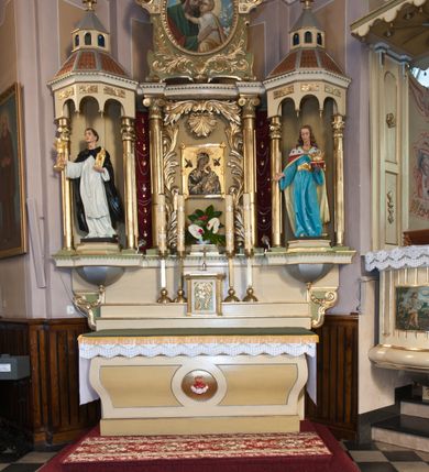 Zdjęcie nr 2: Figura św. Kazimierza Jagiellończyka ustawiona na niskim cokole. Święty ukazany w pozycji stojącej, frontalnie, w lekkim kontrapoście, w lewej dłoni trzyma koronę otwartą ułożoną na czerwonej poduszce, prawą wysuwa do przodu. Twarz szeroka o ostrych rysach, dużych i głęboko osadzonych oczach, długim i szerokim nosie, bez zarostu. Włosy długie, z przedziałkiem pośrodku, opadające bujnymi falami na plecy. Święty jest ubrany w długą, błękitną suknię z szerokimi rękawami, ze złotym obszyciem wokół szyi, spiętą w talii złotym pasem. Spod sukni widoczne złocone trzewiki zakończone kwadratowymi noskami. Na plecy zarzucony długi, czerwony płaszcz, z jasnożółtą podszewką, z gronostajową pelerynką, spięty na piersiach złotą fibulą. Polichromia w odsłoniętych partiach ciała naturalistyczna, detale złocone.