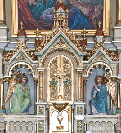 Zdjęcie nr 1: Tabernakulum trójosiowe w formie prostopadłościennej skrzynki z tronem wystawienia, rozbudowanym architektonicznie, ustawionym na niskim cokole dekorowanym arkadowaniem. Pośrodku tron wystawienia flankowany odwróconą parą odrzwi z figurami aniołów, z monogramem IHS w polu na białym tle, zamknięty trójkątnym przyczółkiem, na szczycie kopuła wsparta na wąskich kolumienkach zwieńczona krzyżykiem. W osiach bocznych arkady z figurami klęczących aniołów, zamknięte zwieńczeniem analogicznym jak w osi środkowej. Struktura polichromowana na kolor beżowy, detale na kolor jasnoszary i zielony, ornamentyka i detale złocone. 
