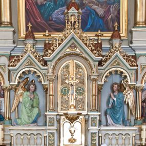 Zdjęcie nr 1: Tabernakulum trójosiowe w formie prostopadłościennej skrzynki z tronem wystawienia, rozbudowanym architektonicznie, ustawionym na niskim cokole dekorowanym arkadowaniem. Pośrodku tron wystawienia flankowany odwróconą parą odrzwi z figurami aniołów, z monogramem IHS w polu na białym tle, zamknięty trójkątnym przyczółkiem, na szczycie kopuła wsparta na wąskich kolumienkach zwieńczona krzyżykiem. W osiach bocznych arkady z figurami klęczących aniołów, zamknięte zwieńczeniem analogicznym jak w osi środkowej. Struktura polichromowana na kolor beżowy, detale na kolor jasnoszary i zielony, ornamentyka i detale złocone. 
