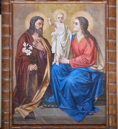 Zdjęcie nr 1: Obraz w kształcie stojącego prostokąta z przedstawieniem Świętej Rodziny. Po prawej stronie siedzi Maria przytrzymując rękami stojące na jej prawym kolanie Dzieciątko Jezus, które lewą dłonią przytrzymuje się jej ramienia, a prawą unosi w geście błogosławieństwa. Maria ma drobną twarz, ukazaną z lewego profilu, prosty i długi nos, duże oczy i delikatnie zaznaczone usta. Brązowe i zwinięte w pukle włosy opadają na jej lewe ramię i plecy. Maria ubrana jest w czerwoną suknię z dekoltem w kształcie litery „V”, ozdobionym złotą bordiurą oraz obszerny niebieski płaszcz z białą podszewką, który opada na kamienny podest ostro załamanymi fałdami. Dzieciątko ubrane jest w białą szatę, luźno zarzuconą na lewe ramię, którą z prawej strony podtrzymuje Maria. Po lewej stronie obrazu stoi św. Józef, zwrócony trzy czwarte w lewo z gałązką białej lilii w prawej dłoni i lewą ręką złożoną na piersi. Twarz podłużna z długim nosem, dużymi oczami, okolona bujną brodą oraz gęstymi, ciemnymi włosami, opadającymi do ramion. Święty ubrany jest w fioletową suknię oraz brązowy płaszcz z jasnobrązową podszewką. Święta Rodzina ukazana została na kamiennym, profilowanym postumencie. Wokół głów postaci zostały umieszczone dekoracyjne koliste nimby, u Dzieciątka Jezus z motywem krzyża, u Marii zdobiony puklowaniem, a wszystkie obficie dekorowane małymi kreseczkami i białymi kropkami. Z tyłu na szarym tle wisi pas złotej tkaniny z motywem małych krzyżyków zamkniętych w podwójnych czwórliściach.