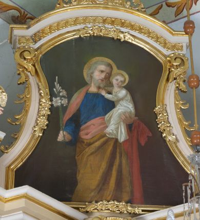 Zdjęcie nr 1: Obraz w kształcie stojącego prostokąta o wklęsło-wypukłych bokach. W centrum kompozycji św. Józef ukazany frontalnie, do wysokości kolan, z Dzieciątkiem Jezus na lewej ręce i gałązką białej lilii w prawej, z głową delikatnie zwróconą w lewo. Święty ubrany jest w błękitną suknię z długimi rękawami, obszytą złotą lamówką oraz jasnobrązowy płaszcz z różową podszewką, założony na plecy i spływający pod prawą ręką na lewe ramię. Twarz owalna, okolona średniej długości, siwą brodą, wzrok skierowany do góry, włosy siwe, zasłaniające uszy. Dzieciątko ukazane w pozycji siedzącej, zwrócone trzy czwarte w prawo, z głową skierowaną na wprost, rękami obejmujące za szyję św. Józefa. Ubrane w długą, białą sukienkę z długimi rękawami. Dłonie św. Józefa i Dzieciątka smukłe, o długich i subtelnych palcach. Wokół głów św. Józefa i Dzieciątka złote, koliste nimby. Tło obrazu jednolite i ciemnozielone.