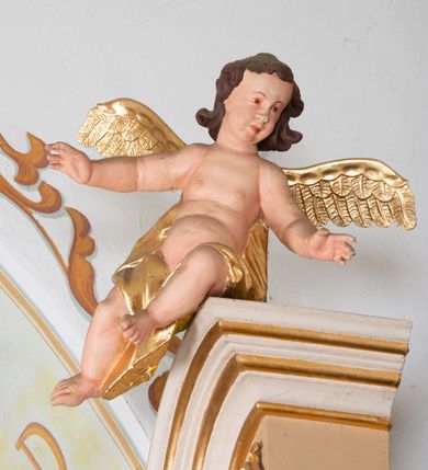 Zdjęcie nr 7: Rzeźby aniołków ukazane są w pozycji siedzącej, zwrócone ku sobie w lustrzanym odbiciu, z głowami uniesionym do góry i z rękami na wysokości piersi, z szeroko rozpostartymi skrzydłami; w pasie przewiązane są złotą chustą. Mają subtelny modelunek ciała. Twarze ich są okrągłe, z pucułowatymi policzkami. Włosy średniej długości, ciemnobrązowe, zasłaniające uszy. Polichromia w odsłoniętych partiach ciała naturalistyczna, skrzydła i chusta złocone. 
