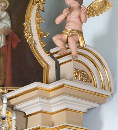 Zdjęcie nr 9: Rzeźby aniołków ukazane są w pozycji siedzącej, zwrócone ku sobie w lustrzanym odbiciu, z głowami uniesionym do góry i z rękami na wysokości piersi, z szeroko rozpostartymi skrzydłami; w pasie przewiązane są złotą chustą. Mają subtelny modelunek ciała. Twarze ich są okrągłe, z pucułowatymi policzkami. Włosy średniej długości, ciemnobrązowe, zasłaniające uszy. Polichromia w odsłoniętych partiach ciała naturalistyczna, skrzydła i chusta złocone. 
