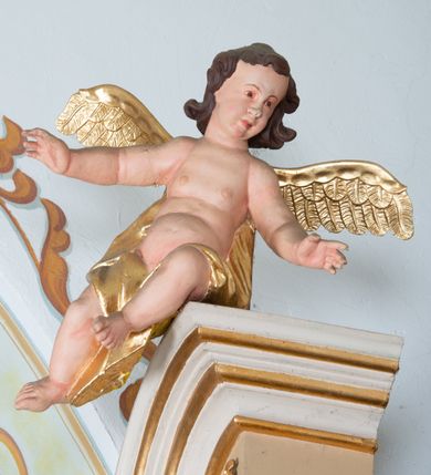 Zdjęcie nr 3: Rzeźby aniołków ukazane są w pozycji siedzącej, zwrócone ku sobie w lustrzanym odbiciu, z głowami uniesionym do góry i z rękami na wysokości piersi, z szeroko rozpostartymi skrzydłami; w pasie przewiązane są złotą chustą. Mają subtelny modelunek ciała. Twarze ich są okrągłe, z pucułowatymi policzkami. Włosy średniej długości, ciemnobrązowe, zasłaniające uszy. Polichromia w odsłoniętych partiach ciała naturalistyczna, skrzydła i chusta złocone. 
