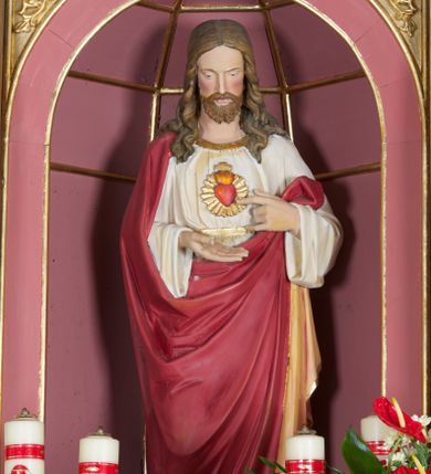 Zdjęcie nr 1: Rzeźba ustawiona na niskim prostopadłościennym postumencie o ściętych narożach. Chrystus ukazany frontalnie, w postawie stojącej, w delikatnym kontrapoście, z prawą nogą ugiętą w kolanie, z głową skierowaną w dół. Jego lewa ręka wskazuje na gorejące serce, zaś prawa, zgięta w łokciu jest wyciągnięta do przodu. Jest ubrany w długą, białą suknię z długimi i szerokimi rękawami, u szyi obwiedzioną złotą lamówką, przewiązaną w talii złotym paskiem oraz czerwony płaszcz z jasnobrązową podszewką, obwiedziony złotą lamówką, założony na prawe ramię, z jedną połą spływającą wzdłuż prawej ręki na lewe przedramię. Na piersi Chrystusa gorejące serce na tle promienistej glorii, z zatkniętym u szczytu małym krzyżykiem. Twarz podłużna z długim i wąskim nosem, małymi oczami, zarumienionymi policzkami, okolona krótką i brązową brodą oraz długimi włosami, z przedziałkiem pośrodku, spływającymi długimi puklami na ramiona i plecy. Polichromia w odsłoniętych partiach ciała naturalistyczna, detale złocone.
