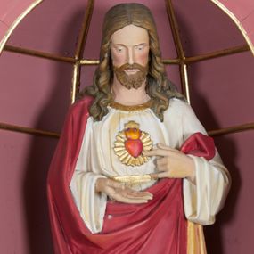 Zdjęcie nr 1: Rzeźba ustawiona na niskim prostopadłościennym postumencie o ściętych narożach. Chrystus ukazany frontalnie, w postawie stojącej, w delikatnym kontrapoście, z prawą nogą ugiętą w kolanie, z głową skierowaną w dół. Jego lewa ręka wskazuje na gorejące serce, zaś prawa, zgięta w łokciu jest wyciągnięta do przodu. Jest ubrany w długą, białą suknię z długimi i szerokimi rękawami, u szyi obwiedzioną złotą lamówką, przewiązaną w talii złotym paskiem oraz czerwony płaszcz z jasnobrązową podszewką, obwiedziony złotą lamówką, założony na prawe ramię, z jedną połą spływającą wzdłuż prawej ręki na lewe przedramię. Na piersi Chrystusa gorejące serce na tle promienistej glorii, z zatkniętym u szczytu małym krzyżykiem. Twarz podłużna z długim i wąskim nosem, małymi oczami, zarumienionymi policzkami, okolona krótką i brązową brodą oraz długimi włosami, z przedziałkiem pośrodku, spływającymi długimi puklami na ramiona i plecy. Polichromia w odsłoniętych partiach ciała naturalistyczna, detale złocone.
