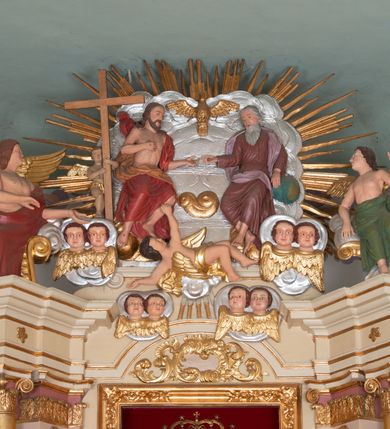 Zdjęcie nr 4: Ołtarz prostopadłościenny, murowany, ustawiony na jednym stopniu. Mensa drewniana, z marmurowym portatylem. W polu antependium rocaille’owy kartusz ujęty parą gałązek palmowych. 

Tabernakulum w formie drewnianej, prostopadłościennej skrzynki na wysokim cokole, o wklęsło-wypukłym rzucie; flankowane parą ustawionych ukośnie kolumienek o kompozytowych kapitelach, dźwigających wyłamane belkowanie. Drzwiczki w formie stojącego prostokąta, obite blachą miedzianą z aplikowanym kielichem z hostią oraz krzyżem na tle promienistej glorii. Nad nimi płaskorzeźbiony, stylizowany ornament rocaille’owy z muszlą palmetową pośrodku. Ścianki boczne dekorowane prostokątnymi płycinami zamkniętymi rocaille’ami z kwiatowymi zwisami. 

Nastawa ołtarzowa architektoniczna, jednokondygnacyjna ze zwieńczeniem, ustawiona na dwustrefowym cokole. Kondygnacja ujęta ustawionymi ukośnie: parą lizen, filarów i dwiema parami kolumn. Podpory dźwigają pełne belkowanie, wyłamane nad osią główną i osiami bocznymi. Podpory ustawione na bazach, o kapitelach kompozytowych; trzony kolumn gładkie, złocone. Na belkowaniu, na wewnętrznej parze podpór siedzące na obłokach figury aniołów. W polu środkowym nisza w formie stojącego prostokąta z obrazem Matki Boskiej Bieńkowskiej, ujęta profilowaną ramą. Nad polem środkowym łuk segmentowy, nadwieszony, zdobiony w polu stylizowanym ornamentem rocaille’owym. Nad łukiem i w zwieńczeniu cztery pary uskrzydlonych główek anielskich na obłokach. Zwieńczenie tworzy grupa rzeźbiarska z przedstawieniem Trójcy Świętej na tle obłoków i promienistej glorii. Filary od wewnątrz dekorowane zwisami utworzonymi z ornamentu rocaille’owego, różanych gałązek, słoneczników, drobnych kwiatów i owoców. Fryz zdobiony pojedynczymi, pięciopłatkowymi kwiatkami. Kondygnacja ujęta uszami w kształcie drapowanej kotary, obwiedzionej złotą lamówką, podtrzymywanej przez parę puttów. Putta ustawione frontalnie, ze skrzyżowanymi nogami, z jedną ręką uniesioną do góry, drugą spuszczoną w dół, z jednym skrzydłem od strony wewnętrznej, z pasem tkaniny spływającym z ramienia na biodra. Putta rzeźbione miękko; twarze owalne, pucułowate, z małymi oczami i ustami; włosy jasne, kędzierzawe. 

Struktura polichromowana w kolorze beżowym z różowymi detalami; kolumny, profile i ornamentyka złocone i srebrzone.
