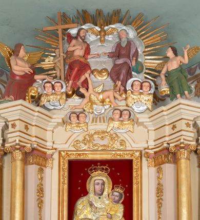 Zdjęcie nr 1: Zwieńczenie ołtarza w formie rzeźbiarskiej grupy z przedstawieniem Trójcy Świętej na tle obłoków i promienistej glorii; figury ścięte z tyłu. Po prawej stronie figura Boga Ojca, zwróconego delikatnie w prawo, w pozycji siedzącej, ze skrzyżowanymi nogami, z lewą ręką wspartą na globie ziemskim, a z prawą wyciągniętą do Chrystusa. Po lewej stronie figura Chrystusa zwróconego trzy czwarte w lewo, w pozycji siedzącej, z prawą ręką złożoną na piersi, a z lewą wyciągniętą do Boga Ojca. Nad nimi gołębica Ducha Świętego z rozpostartymi szeroko skrzydłami. Bóg Ojciec ubrany w ciemnoróżową suknię z długimi rękawami, delikatnie drapowaną oraz jasnofioletowy płaszcz zarzucony na plecy i spływający jedną połą na biodra. Chrystus ubrany w czerwony płaszcz, z jasnobrązową podszewką, odsłaniający nagi tors. Twarz Boga Ojca owalna, z długim i wąskim nosem, z zarumienionymi policzkami, okolona długimi i siwymi włosami, spływającymi na plecy oraz długą brodą. Chrystus o twarzy podłużnej, z długim i wąskim nosem, okolonej krótką, ciemnobrązową brodą i długimi włosami spływającymi na plecy i ramiona. Na ciele Chrystusa widoczne ślady męki. Po lewej stronie, za plecami Chrystusa znajduje się niewielkie putto ukazane frontalnie, z prawą nogą wysuniętą do przodu, w rękach trzymające wysoki krzyż. Poniżej, pośrodku putto na obłoku, ukazane w pozycji leżącej, z prawego profilu, z rękami wzniesionymi do góry oraz cztery pary uskrzydlonych główek anielskich na tle obłoków. 

Polichromia w odsłoniętych partiach ciała naturalistyczna, obłoki oraz gloria srebrzone i złocone.
