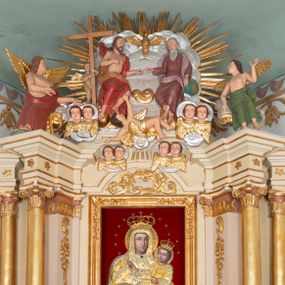 Zdjęcie nr 1: Zwieńczenie ołtarza w formie rzeźbiarskiej grupy z przedstawieniem Trójcy Świętej na tle obłoków i promienistej glorii; figury ścięte z tyłu. Po prawej stronie figura Boga Ojca, zwróconego delikatnie w prawo, w pozycji siedzącej, ze skrzyżowanymi nogami, z lewą ręką wspartą na globie ziemskim, a z prawą wyciągniętą do Chrystusa. Po lewej stronie figura Chrystusa zwróconego trzy czwarte w lewo, w pozycji siedzącej, z prawą ręką złożoną na piersi, a z lewą wyciągniętą do Boga Ojca. Nad nimi gołębica Ducha Świętego z rozpostartymi szeroko skrzydłami. Bóg Ojciec ubrany w ciemnoróżową suknię z długimi rękawami, delikatnie drapowaną oraz jasnofioletowy płaszcz zarzucony na plecy i spływający jedną połą na biodra. Chrystus ubrany w czerwony płaszcz, z jasnobrązową podszewką, odsłaniający nagi tors. Twarz Boga Ojca owalna, z długim i wąskim nosem, z zarumienionymi policzkami, okolona długimi i siwymi włosami, spływającymi na plecy oraz długą brodą. Chrystus o twarzy podłużnej, z długim i wąskim nosem, okolonej krótką, ciemnobrązową brodą i długimi włosami spływającymi na plecy i ramiona. Na ciele Chrystusa widoczne ślady męki. Po lewej stronie, za plecami Chrystusa znajduje się niewielkie putto ukazane frontalnie, z prawą nogą wysuniętą do przodu, w rękach trzymające wysoki krzyż. Poniżej, pośrodku putto na obłoku, ukazane w pozycji leżącej, z prawego profilu, z rękami wzniesionymi do góry oraz cztery pary uskrzydlonych główek anielskich na tle obłoków. 

Polichromia w odsłoniętych partiach ciała naturalistyczna, obłoki oraz gloria srebrzone i złocone.
