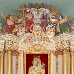 Zdjęcie nr 1: Zwieńczenie ołtarza w formie rzeźbiarskiej grupy z przedstawieniem Trójcy Świętej na tle obłoków i promienistej glorii; figury ścięte z tyłu. Po prawej stronie figura Boga Ojca, zwróconego delikatnie w prawo, w pozycji siedzącej, ze skrzyżowanymi nogami, z lewą ręką wspartą na globie ziemskim, a z prawą wyciągniętą do Chrystusa. Po lewej stronie figura Chrystusa zwróconego trzy czwarte w lewo, w pozycji siedzącej, z prawą ręką złożoną na piersi, a z lewą wyciągniętą do Boga Ojca. Nad nimi gołębica Ducha Świętego z rozpostartymi szeroko skrzydłami. Bóg Ojciec ubrany w ciemnoróżową suknię z długimi rękawami, delikatnie drapowaną oraz jasnofioletowy płaszcz zarzucony na plecy i spływający jedną połą na biodra. Chrystus ubrany w czerwony płaszcz, z jasnobrązową podszewką, odsłaniający nagi tors. Twarz Boga Ojca owalna, z długim i wąskim nosem, z zarumienionymi policzkami, okolona długimi i siwymi włosami, spływającymi na plecy oraz długą brodą. Chrystus o twarzy podłużnej, z długim i wąskim nosem, okolonej krótką, ciemnobrązową brodą i długimi włosami spływającymi na plecy i ramiona. Na ciele Chrystusa widoczne ślady męki. Po lewej stronie, za plecami Chrystusa znajduje się niewielkie putto ukazane frontalnie, z prawą nogą wysuniętą do przodu, w rękach trzymające wysoki krzyż. Poniżej, pośrodku putto na obłoku, ukazane w pozycji leżącej, z prawego profilu, z rękami wzniesionymi do góry oraz cztery pary uskrzydlonych główek anielskich na tle obłoków. 

Polichromia w odsłoniętych partiach ciała naturalistyczna, obłoki oraz gloria srebrzone i złocone.
