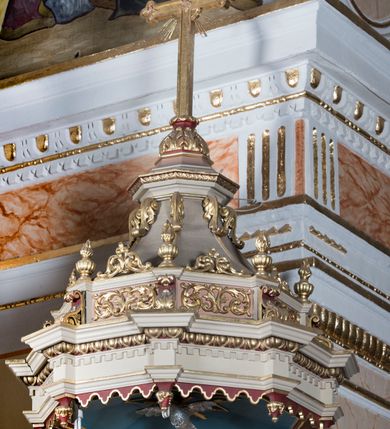 Zdjęcie nr 5: Ambona przyścienna, wisząca z dostępem od strony wewnętrznej klatki schodowej usytuowanej w murze kościoła. Kosz jest na planie ośmioboku z cokołem i podwieszeniem w formie stylizowanego, odwróconego stożka zakończonego szyszką. Po lewej stronie przylega do niego ganek na rzucie czworoboku. Naroża kosza i ganku ujęte są kolumienkami, których trzony w dolnej partii zdobią liście akantu i rozetki, wyżej obiega je para pierścieni, pomiędzy którymi naprzemiennie są kaboszony i rozetki, natomiast górna część jest kanelurowana. Na kompozytowych kapitelach wsparte jest belkowanie parapetu. Zaplecek ambony ujmuje para osadzonych na cokołach kolumienek w dolnej partii zdobionych motywem floralnym, w górnej kanelurowaniem, wpierających wyłamujące się ponad nimi, przerwane belkowanie. Baldachim jest na rzucie ośmioboku. Jego krawędź obiega wyłamujące się na narożach belkowanie. Zwieńczenie ma formę osadzonego na niewysokim cokole stożka o lekko wklęsłych ściankach, nakrytego wklęsło-wypukłym hełmem na rzucie ośmioboku, na którym osadzony jest złocony krzyż o trójlistnych zakończeniach ramion oraz z kręgiem obłoków i wiązkami promieni na ich przecięciu. Płyciny podwieszenia kosza zdobią liście akantu. Na cokole kosza i ganku są girlandy z kwiatami róż, natomiast na fryzie akantowa wić. Ścianki ganku artykułują prostokątne płyciny natomiast kosza – w kształcie stojącego prostokąta zamkniętego łukiem półkolistym. Ujęte są profilowaną, złoconą ramą i wypełnione dekoracyjnym motywem wazonu z kompozycją liści akantu o symetrycznym układzie. W polu zaplecka znajduje się płycina w kształcie stojącego prostokąta zamknięta półkolistym łukiem nadwieszonym z górnymi narożnikami zaakcentowanymi przez wklęsłe łuki odcinkowe. Ujęta jest profilowaną, złoconą ramą. Wypełnia ją motyw dekoracyjny złożony z ukazanych na tle krzyżującego się kwiatu lilii i gałązki palmowej, skrzyżowanych kotwicy i krzyża, z sercem gorejącym pośrodku, dodatkowo u dołu kompozycji jest para kwiatów róży, a u góry wstążka. W podniebiu baldachimu znajduje się gołębica Ducha Świętego na tle promienistej glorii. Dolną krawędź baldachimu obiega motyw lambrekinu, belkowanie zdobi ornament kostkowy i wole oczy. Dekorację cokołu zwieńczenia stanowią motywy akantowe, artykułujące naroża akantowe woluty oraz wazony. Natomiast górną część zwieńczenia i hełm zdobią akantowe liście. Struktura ambony utrzymana jest w bieli, czerwieni, błękicie i jasnym różu, detal architektoniczny i ornamentyka są złocone.  