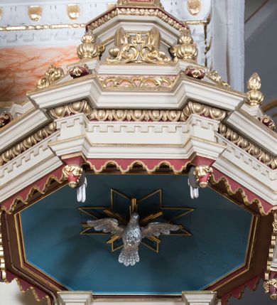 Zdjęcie nr 4: Ambona przyścienna, wisząca z dostępem od strony wewnętrznej klatki schodowej usytuowanej w murze kościoła. Kosz jest na planie ośmioboku z cokołem i podwieszeniem w formie stylizowanego, odwróconego stożka zakończonego szyszką. Po lewej stronie przylega do niego ganek na rzucie czworoboku. Naroża kosza i ganku ujęte są kolumienkami, których trzony w dolnej partii zdobią liście akantu i rozetki, wyżej obiega je para pierścieni, pomiędzy którymi naprzemiennie są kaboszony i rozetki, natomiast górna część jest kanelurowana. Na kompozytowych kapitelach wsparte jest belkowanie parapetu. Zaplecek ambony ujmuje para osadzonych na cokołach kolumienek w dolnej partii zdobionych motywem floralnym, w górnej kanelurowaniem, wpierających wyłamujące się ponad nimi, przerwane belkowanie. Baldachim jest na rzucie ośmioboku. Jego krawędź obiega wyłamujące się na narożach belkowanie. Zwieńczenie ma formę osadzonego na niewysokim cokole stożka o lekko wklęsłych ściankach, nakrytego wklęsło-wypukłym hełmem na rzucie ośmioboku, na którym osadzony jest złocony krzyż o trójlistnych zakończeniach ramion oraz z kręgiem obłoków i wiązkami promieni na ich przecięciu. Płyciny podwieszenia kosza zdobią liście akantu. Na cokole kosza i ganku są girlandy z kwiatami róż, natomiast na fryzie akantowa wić. Ścianki ganku artykułują prostokątne płyciny natomiast kosza – w kształcie stojącego prostokąta zamkniętego łukiem półkolistym. Ujęte są profilowaną, złoconą ramą i wypełnione dekoracyjnym motywem wazonu z kompozycją liści akantu o symetrycznym układzie. W polu zaplecka znajduje się płycina w kształcie stojącego prostokąta zamknięta półkolistym łukiem nadwieszonym z górnymi narożnikami zaakcentowanymi przez wklęsłe łuki odcinkowe. Ujęta jest profilowaną, złoconą ramą. Wypełnia ją motyw dekoracyjny złożony z ukazanych na tle krzyżującego się kwiatu lilii i gałązki palmowej, skrzyżowanych kotwicy i krzyża, z sercem gorejącym pośrodku, dodatkowo u dołu kompozycji jest para kwiatów róży, a u góry wstążka. W podniebiu baldachimu znajduje się gołębica Ducha Świętego na tle promienistej glorii. Dolną krawędź baldachimu obiega motyw lambrekinu, belkowanie zdobi ornament kostkowy i wole oczy. Dekorację cokołu zwieńczenia stanowią motywy akantowe, artykułujące naroża akantowe woluty oraz wazony. Natomiast górną część zwieńczenia i hełm zdobią akantowe liście. Struktura ambony utrzymana jest w bieli, czerwieni, błękicie i jasnym różu, detal architektoniczny i ornamentyka są złocone.  