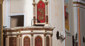 Zdjęcie nr 1: Ambona przyścienna, wisząca z dostępem od strony wewnętrznej klatki schodowej usytuowanej w murze kościoła. Kosz jest na planie ośmioboku z cokołem i podwieszeniem w formie stylizowanego, odwróconego stożka zakończonego szyszką. Po lewej stronie przylega do niego ganek na rzucie czworoboku. Naroża kosza i ganku ujęte są kolumienkami, których trzony w dolnej partii zdobią liście akantu i rozetki, wyżej obiega je para pierścieni, pomiędzy którymi naprzemiennie są kaboszony i rozetki, natomiast górna część jest kanelurowana. Na kompozytowych kapitelach wsparte jest belkowanie parapetu. Zaplecek ambony ujmuje para osadzonych na cokołach kolumienek w dolnej partii zdobionych motywem floralnym, w górnej kanelurowaniem, wpierających wyłamujące się ponad nimi, przerwane belkowanie. Baldachim jest na rzucie ośmioboku. Jego krawędź obiega wyłamujące się na narożach belkowanie. Zwieńczenie ma formę osadzonego na niewysokim cokole stożka o lekko wklęsłych ściankach, nakrytego wklęsło-wypukłym hełmem na rzucie ośmioboku, na którym osadzony jest złocony krzyż o trójlistnych zakończeniach ramion oraz z kręgiem obłoków i wiązkami promieni na ich przecięciu. Płyciny podwieszenia kosza zdobią liście akantu. Na cokole kosza i ganku są girlandy z kwiatami róż, natomiast na fryzie akantowa wić. Ścianki ganku artykułują prostokątne płyciny natomiast kosza – w kształcie stojącego prostokąta zamkniętego łukiem półkolistym. Ujęte są profilowaną, złoconą ramą i wypełnione dekoracyjnym motywem wazonu z kompozycją liści akantu o symetrycznym układzie. W polu zaplecka znajduje się płycina w kształcie stojącego prostokąta zamknięta półkolistym łukiem nadwieszonym z górnymi narożnikami zaakcentowanymi przez wklęsłe łuki odcinkowe. Ujęta jest profilowaną, złoconą ramą. Wypełnia ją motyw dekoracyjny złożony z ukazanych na tle krzyżującego się kwiatu lilii i gałązki palmowej, skrzyżowanych kotwicy i krzyża, z sercem gorejącym pośrodku, dodatkowo u dołu kompozycji jest para kwiatów róży, a u góry wstążka. W podniebiu baldachimu znajduje się gołębica Ducha Świętego na tle promienistej glorii. Dolną krawędź baldachimu obiega motyw lambrekinu, belkowanie zdobi ornament kostkowy i wole oczy. Dekorację cokołu zwieńczenia stanowią motywy akantowe, artykułujące naroża akantowe woluty oraz wazony. Natomiast górną część zwieńczenia i hełm zdobią akantowe liście. Struktura ambony utrzymana jest w bieli, czerwieni, błękicie i jasnym różu, detal architektoniczny i ornamentyka są złocone.  