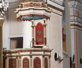 Zdjęcie nr 1: Ambona przyścienna, wisząca z dostępem od strony wewnętrznej klatki schodowej usytuowanej w murze kościoła. Kosz jest na planie ośmioboku z cokołem i podwieszeniem w formie stylizowanego, odwróconego stożka zakończonego szyszką. Po lewej stronie przylega do niego ganek na rzucie czworoboku. Naroża kosza i ganku ujęte są kolumienkami, których trzony w dolnej partii zdobią liście akantu i rozetki, wyżej obiega je para pierścieni, pomiędzy którymi naprzemiennie są kaboszony i rozetki, natomiast górna część jest kanelurowana. Na kompozytowych kapitelach wsparte jest belkowanie parapetu. Zaplecek ambony ujmuje para osadzonych na cokołach kolumienek w dolnej partii zdobionych motywem floralnym, w górnej kanelurowaniem, wpierających wyłamujące się ponad nimi, przerwane belkowanie. Baldachim jest na rzucie ośmioboku. Jego krawędź obiega wyłamujące się na narożach belkowanie. Zwieńczenie ma formę osadzonego na niewysokim cokole stożka o lekko wklęsłych ściankach, nakrytego wklęsło-wypukłym hełmem na rzucie ośmioboku, na którym osadzony jest złocony krzyż o trójlistnych zakończeniach ramion oraz z kręgiem obłoków i wiązkami promieni na ich przecięciu. Płyciny podwieszenia kosza zdobią liście akantu. Na cokole kosza i ganku są girlandy z kwiatami róż, natomiast na fryzie akantowa wić. Ścianki ganku artykułują prostokątne płyciny natomiast kosza – w kształcie stojącego prostokąta zamkniętego łukiem półkolistym. Ujęte są profilowaną, złoconą ramą i wypełnione dekoracyjnym motywem wazonu z kompozycją liści akantu o symetrycznym układzie. W polu zaplecka znajduje się płycina w kształcie stojącego prostokąta zamknięta półkolistym łukiem nadwieszonym z górnymi narożnikami zaakcentowanymi przez wklęsłe łuki odcinkowe. Ujęta jest profilowaną, złoconą ramą. Wypełnia ją motyw dekoracyjny złożony z ukazanych na tle krzyżującego się kwiatu lilii i gałązki palmowej, skrzyżowanych kotwicy i krzyża, z sercem gorejącym pośrodku, dodatkowo u dołu kompozycji jest para kwiatów róży, a u góry wstążka. W podniebiu baldachimu znajduje się gołębica Ducha Świętego na tle promienistej glorii. Dolną krawędź baldachimu obiega motyw lambrekinu, belkowanie zdobi ornament kostkowy i wole oczy. Dekorację cokołu zwieńczenia stanowią motywy akantowe, artykułujące naroża akantowe woluty oraz wazony. Natomiast górną część zwieńczenia i hełm zdobią akantowe liście. Struktura ambony utrzymana jest w bieli, czerwieni, błękicie i jasnym różu, detal architektoniczny i ornamentyka są złocone.  