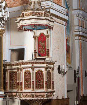 Zdjęcie nr 1: Ambona przyścienna, wisząca z dostępem od strony wewnętrznej klatki schodowej usytuowanej w murze kościoła. Kosz jest na planie ośmioboku z cokołem i podwieszeniem w formie stylizowanego, odwróconego stożka zakończonego szyszką. Po lewej stronie przylega do niego ganek na rzucie czworoboku. Naroża kosza i ganku ujęte są kolumienkami, których trzony w dolnej partii zdobią liście akantu i rozetki, wyżej obiega je para pierścieni, pomiędzy którymi naprzemiennie są kaboszony i rozetki, natomiast górna część jest kanelurowana. Na kompozytowych kapitelach wsparte jest belkowanie parapetu. Zaplecek ambony ujmuje para osadzonych na cokołach kolumienek w dolnej partii zdobionych motywem floralnym, w górnej kanelurowaniem, wpierających wyłamujące się ponad nimi, przerwane belkowanie. Baldachim jest na rzucie ośmioboku. Jego krawędź obiega wyłamujące się na narożach belkowanie. Zwieńczenie ma formę osadzonego na niewysokim cokole stożka o lekko wklęsłych ściankach, nakrytego wklęsło-wypukłym hełmem na rzucie ośmioboku, na którym osadzony jest złocony krzyż o trójlistnych zakończeniach ramion oraz z kręgiem obłoków i wiązkami promieni na ich przecięciu. Płyciny podwieszenia kosza zdobią liście akantu. Na cokole kosza i ganku są girlandy z kwiatami róż, natomiast na fryzie akantowa wić. Ścianki ganku artykułują prostokątne płyciny natomiast kosza – w kształcie stojącego prostokąta zamkniętego łukiem półkolistym. Ujęte są profilowaną, złoconą ramą i wypełnione dekoracyjnym motywem wazonu z kompozycją liści akantu o symetrycznym układzie. W polu zaplecka znajduje się płycina w kształcie stojącego prostokąta zamknięta półkolistym łukiem nadwieszonym z górnymi narożnikami zaakcentowanymi przez wklęsłe łuki odcinkowe. Ujęta jest profilowaną, złoconą ramą. Wypełnia ją motyw dekoracyjny złożony z ukazanych na tle krzyżującego się kwiatu lilii i gałązki palmowej, skrzyżowanych kotwicy i krzyża, z sercem gorejącym pośrodku, dodatkowo u dołu kompozycji jest para kwiatów róży, a u góry wstążka. W podniebiu baldachimu znajduje się gołębica Ducha Świętego na tle promienistej glorii. Dolną krawędź baldachimu obiega motyw lambrekinu, belkowanie zdobi ornament kostkowy i wole oczy. Dekorację cokołu zwieńczenia stanowią motywy akantowe, artykułujące naroża akantowe woluty oraz wazony. Natomiast górną część zwieńczenia i hełm zdobią akantowe liście. Struktura ambony utrzymana jest w bieli, czerwieni, błękicie i jasnym różu, detal architektoniczny i ornamentyka są złocone.  