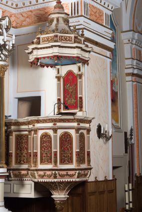 Zdjęcie nr 1: Ambona przyścienna, wisząca z dostępem od strony wewnętrznej klatki schodowej usytuowanej w murze kościoła. Kosz jest na planie ośmioboku z cokołem i podwieszeniem w formie stylizowanego, odwróconego stożka zakończonego szyszką. Po lewej stronie przylega do niego ganek na rzucie czworoboku. Naroża kosza i ganku ujęte są kolumienkami, których trzony w dolnej partii zdobią liście akantu i rozetki, wyżej obiega je para pierścieni, pomiędzy którymi naprzemiennie są kaboszony i rozetki, natomiast górna część jest kanelurowana. Na kompozytowych kapitelach wsparte jest belkowanie parapetu. Zaplecek ambony ujmuje para osadzonych na cokołach kolumienek w dolnej partii zdobionych motywem floralnym, w górnej kanelurowaniem, wpierających wyłamujące się ponad nimi, przerwane belkowanie. Baldachim jest na rzucie ośmioboku. Jego krawędź obiega wyłamujące się na narożach belkowanie. Zwieńczenie ma formę osadzonego na niewysokim cokole stożka o lekko wklęsłych ściankach, nakrytego wklęsło-wypukłym hełmem na rzucie ośmioboku, na którym osadzony jest złocony krzyż o trójlistnych zakończeniach ramion oraz z kręgiem obłoków i wiązkami promieni na ich przecięciu. Płyciny podwieszenia kosza zdobią liście akantu. Na cokole kosza i ganku są girlandy z kwiatami róż, natomiast na fryzie akantowa wić. Ścianki ganku artykułują prostokątne płyciny natomiast kosza – w kształcie stojącego prostokąta zamkniętego łukiem półkolistym. Ujęte są profilowaną, złoconą ramą i wypełnione dekoracyjnym motywem wazonu z kompozycją liści akantu o symetrycznym układzie. W polu zaplecka znajduje się płycina w kształcie stojącego prostokąta zamknięta półkolistym łukiem nadwieszonym z górnymi narożnikami zaakcentowanymi przez wklęsłe łuki odcinkowe. Ujęta jest profilowaną, złoconą ramą. Wypełnia ją motyw dekoracyjny złożony z ukazanych na tle krzyżującego się kwiatu lilii i gałązki palmowej, skrzyżowanych kotwicy i krzyża, z sercem gorejącym pośrodku, dodatkowo u dołu kompozycji jest para kwiatów róży, a u góry wstążka. W podniebiu baldachimu znajduje się gołębica Ducha Świętego na tle promienistej glorii. Dolną krawędź baldachimu obiega motyw lambrekinu, belkowanie zdobi ornament kostkowy i wole oczy. Dekorację cokołu zwieńczenia stanowią motywy akantowe, artykułujące naroża akantowe woluty oraz wazony. Natomiast górną część zwieńczenia i hełm zdobią akantowe liście. Struktura ambony utrzymana jest w bieli, czerwieni, błękicie i jasnym różu, detal architektoniczny i ornamentyka są złocone.  