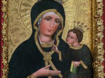 Zdjęcie nr 1: Obraz w kształcie stojącego prostokąta z półpostaciowym przedstawieniem Madonny podtrzymującej lewą ręką Dzieciątko Jezus. Maria ukazana jest na wprost, lekko przechyloną w lewo głowę charakteryzuje owalna, pociągła twarz o wyrazistych oczach z nieznacznie przymkniętymi powiekami, długim, prostym nosie, niewielkich ustach o opadających kącikach, wyraźnie zaróżowionych policzkach oraz subtelnie zaznaczonym podbródku. Maria ubrana jest w ciemnogranatową suknię z szerokimi, dekoracyjnymi, złoconymi obszyciami dekoltu i wąskich mankietów. Na jej głowie jest biała, półprzejrzysta chusta widoczna spod osłaniającego całą sylwetkę ciemnogranatowego płaszcza o bordowej podszewce i szerokiej, złoconej, dekoracyjnej lamówce, spiętego na piersiach dużą, złoconą broszą w kształcie rombu z czterema kółkami przylegającymi do jego lekko wklęsłych boków. Prawą ręką o smukłej, wyprostowanej dłoni Maria wskazuje na trzymane na lewej ręce siedzące Dzieciątko. Zwrócony w trzech czwartych w prawo Jezus lekko pochyla się do przodu. Pociągłą, o trójkątnym obrysie szczęki twarz charakteryzują duże, wyraziste oczy o nieznacznie przymkniętych powiekach, długi, prosty nos oraz niewielkie usta. Krótkie, brązowe, skręcone włosy opadają parą pukli na czoło Dzieciątka. Ubrane jest ono w długą, ciemnogranatową suknię z szerokimi, dekoracyjnymi, złoconymi lamówkami, spod której widoczne są bose stopy. Jezus prawą dłoń unosi w geście błogosławieństwa, lewą trzyma wspartą na kolanach zamkniętą księgę w zielonej oprawie. Głowy postaci okalają ryte w gruncie podobrazia nimby z motywami eliptycznych promieni ujętych wąskim okręgiem, dodatkowo wieńczą je złocone, kameryzowane korony otwarte. Złocone tło przedstawienia zdobi grawerowana wić roślinna. Obraz ujęty jest złoconą ramą w formie ażurowego układu liści akantu.      