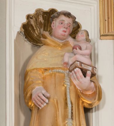 Zdjęcie nr 1: Całopostaciowa, drążona z tyłu figura św. Antoniego Padewskiego stojącego w układzie frontalnym, w nieznacznym kontrapoście, na niewielkim, prostopadłościennym postumencie. Przechyloną lekko w prawo głowę charakteryzuje owalna twarz o pełnych, sumarycznie opracowanych rysach z szeroko rozstawionymi oczami oraz z zarumienionymi policzkami, zaokrąglonym czubkiem nosa i wydatnym podbródkiem. Krótkie, brązowe, skręcone włosy z wyciętą tonsurą odsłaniają duże uszy. Święty ubrany jest w złocony habit franciszkański przewiązany zwisającym sznurem i sandały. Prawa ręka jest opuszczona wzdłuż ciała, lewą podtrzymuje zamkniętą księgę, na której siedzi zwrócone profilem w lewo nagie Dzieciątko Jezus, prawą ręką dotykające twarzy świętego, w lewej trzymające kulę symbolizująca glob ziemski.