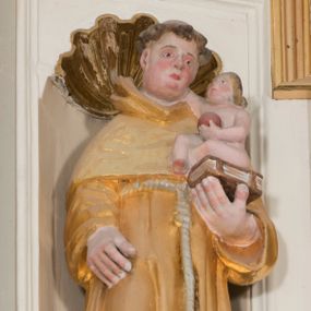 Zdjęcie nr 1: Całopostaciowa, drążona z tyłu figura św. Antoniego Padewskiego stojącego w układzie frontalnym, w nieznacznym kontrapoście, na niewielkim, prostopadłościennym postumencie. Przechyloną lekko w prawo głowę charakteryzuje owalna twarz o pełnych, sumarycznie opracowanych rysach z szeroko rozstawionymi oczami oraz z zarumienionymi policzkami, zaokrąglonym czubkiem nosa i wydatnym podbródkiem. Krótkie, brązowe, skręcone włosy z wyciętą tonsurą odsłaniają duże uszy. Święty ubrany jest w złocony habit franciszkański przewiązany zwisającym sznurem i sandały. Prawa ręka jest opuszczona wzdłuż ciała, lewą podtrzymuje zamkniętą księgę, na której siedzi zwrócone profilem w lewo nagie Dzieciątko Jezus, prawą ręką dotykające twarzy świętego, w lewej trzymające kulę symbolizująca glob ziemski.