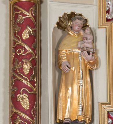 Zdjęcie nr 2: Całopostaciowa, drążona z tyłu figura św. Antoniego Padewskiego stojącego w układzie frontalnym, w nieznacznym kontrapoście, na niewielkim, prostopadłościennym postumencie. Przechyloną lekko w prawo głowę charakteryzuje owalna twarz o pełnych, sumarycznie opracowanych rysach z szeroko rozstawionymi oczami oraz z zarumienionymi policzkami, zaokrąglonym czubkiem nosa i wydatnym podbródkiem. Krótkie, brązowe, skręcone włosy z wyciętą tonsurą odsłaniają duże uszy. Święty ubrany jest w złocony habit franciszkański przewiązany zwisającym sznurem i sandały. Prawa ręka jest opuszczona wzdłuż ciała, lewą podtrzymuje zamkniętą księgę, na której siedzi zwrócone profilem w lewo nagie Dzieciątko Jezus, prawą ręką dotykające twarzy świętego, w lewej trzymające kulę symbolizująca glob ziemski.